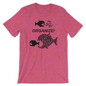 May include: A pink t-shirt with a black graphic of two fish. The smaller fish is being swallowed by the larger fish, which is made up of many smaller fish. The text "ORGANIZE!" is printed above the fish.