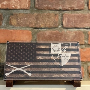 Può includere: Targa in legno raffigurante la bandiera americana con dettagli incisi. Presenta le stelle e le strisce della bandiera, un emblema militare e fucili incrociati. La targa è montata su un supporto in legno, davanti a un muro di mattoni.