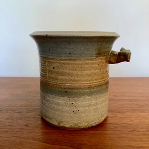 May include: A brown and grey ceramic vase with a unique handle. The vase has a speckled glaze and a striped design.