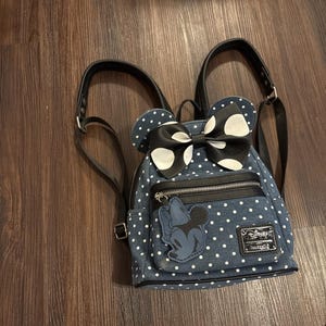 Puede incluir: Una mochila de mezclilla con lunares blancos, con un gran lazo negro con lunares blancos y una aplicación de Minnie Mouse. La mochila tiene correas negras y un cierre de cremallera. Una etiqueta de Disney Loungefly es visible.