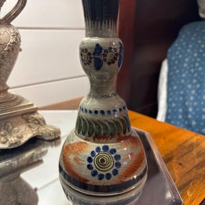 May include: A ceramic candlestick holder with a flared base and a narrow neck. The piece features a light gray glaze decorated with hand-painted floral motifs in blue, green, and orange, including a central spiral pattern and dark blue rim accents.