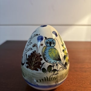 May include: A hand-painted ceramic egg-shaped figurine featuring a detailed owl design. The owl has blue and yellow feathers, perched on a green and brown landscape with floral accents. The base is a light blue and white stripe.