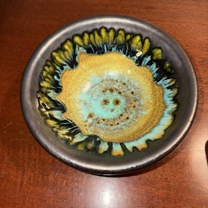 May include: A small, round ceramic bowl with a dark gray exterior and a vibrant interior. The interior features a central gold-colored design surrounded by turquoise and green accents. The bowl's rim is dark gray, creating a striking contrast.