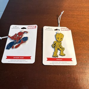 Könnte beinhalten: Zwei Marvel-Weihnachtsbaumschmuck. Einer zeigt Spider-Man in Rot, Blau und Schwarz. Der andere ist Groot in Gelb. Beide befinden sich auf weißen Karten mit dem Marvel-Logo und dem Text "Christbaumschmuck".