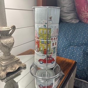 Può includere: Un set di tre tazze cilindriche in ceramica impilate verticalmente in un supporto in filo metallico color argento. Le tazze presentano un'illustrazione continua di un villaggio invernale con case colorate e un'auto rossa su una strada.