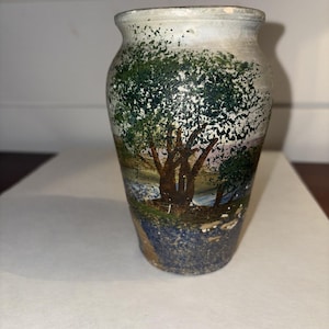 Peut inclure: Petit vase en céramique peint à la main, représentant un paysage. Le vase présente un arbre avec des feuilles vertes et un tronc brun, sur fond bleu et blanc. Le vase a un bord blanc et une surface légèrement texturée.