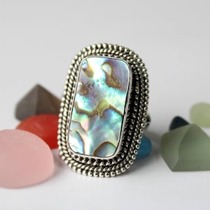 May include: A silver ring with a large, rectangular abalone shell inlay. The ring has a beaded edge and is set against a white background.
