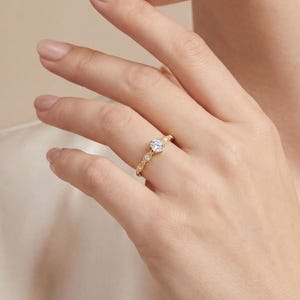 以下が含まれることがあります： 中央に丸い透明な宝石、バンドに沿って小さな透明な宝石が付いたゴールドリング。 リングは人の指にあり、手と腕が見えます。 リングはジュエリーです。