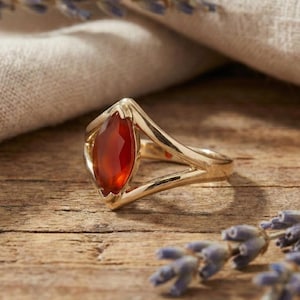 May include: A gold ring with a marquise-cut, reddish-orange gemstone. The ring has a split shank design and is set against a wooden surface with sprigs of lavender and a linen cloth in the background. The ring's setting has a scalloped edge.