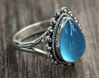 May include: A silver ring with a teardrop-shaped, light blue gemstone. The gemstone is surrounded by a beaded border and set in a detailed silver band. The ring is displayed on a wooden surface.