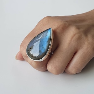 May include: A large, teardrop-shaped ring featuring a polished labradorite stone. The stone displays iridescent blue and gray hues, set in a silver-colored metal band. The ring is shown on a hand against a white background.