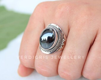 Puede incluir: Un anillo de plata con una piedra negra ovalada grande y pulida. El anillo tiene una banda detallada y texturizada con un diseño en forma de hoja alrededor de la piedra. El texto "VERDIGRIS JEWELLERY" es visible.