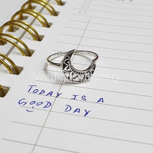 May include: A silver ring with a crescent moon design. The moon is open and has a delicate filigree pattern. The ring is on a white lined notebook with the text "Today is a good day" written in blue ink.