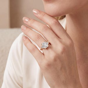 May include: A gold ring with a large, rectangular, emerald-cut center stone and two smaller, trapezoid-shaped side stones. The ring is set on a gold band and is displayed on a person's hand.