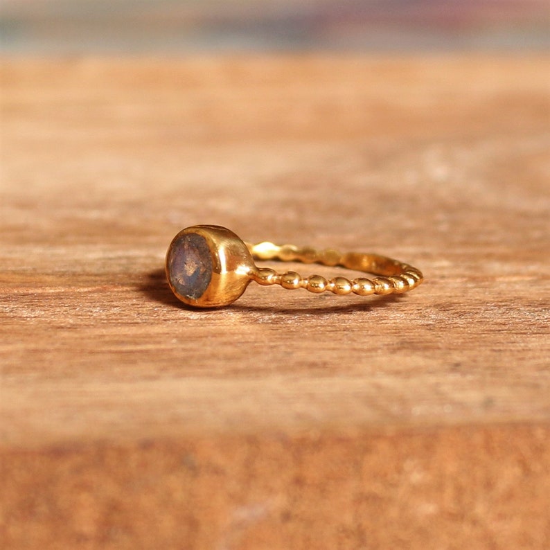 May include: A gold ring with a round, iridescent gemstone set in a bezel setting. The band is made of small, round beads.