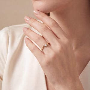 以下が含まれることがあります： 小さな丸い透明な宝石が付いたゴールドリング。リングは繊細な渦巻き模様です。リングは人の指にあります。その人は白いトップスを着ています。