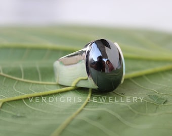 Puede incluir: Un anillo de plata con una gran piedra preciosa negra redonda. El anillo está colocado sobre una hoja verde.