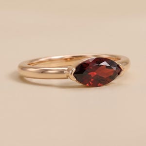 May include: A polished rose gold ring featuring a single marquise-cut deep red garnet gemstone set horizontally. The band is smooth and rounded, showcasing a clean metal finish that complements the faceted dark red stone at the center of the piece.
