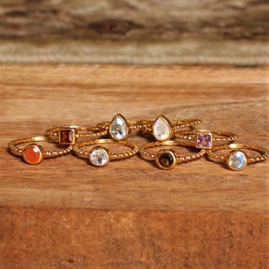 May include: A set of eight gold rings with different colored gemstones. The rings are stacked on top of each other and are arranged in a semi-circle. The gemstones are all different shapes and sizes. The rings are made of gold and have a beaded design.