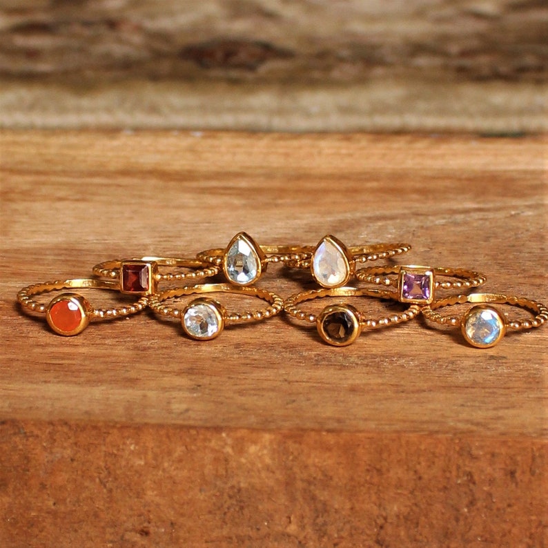 May include: A set of eight gold rings with different colored gemstones. The rings are stacked on top of each other and are arranged in a semi-circle. The gemstones are all different shapes and sizes. The rings are made of gold and have a beaded design.