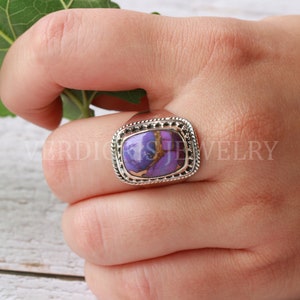 May include: A silver ring with a purple and brown gemstone set in a square bezel. The ring is on a person's finger.