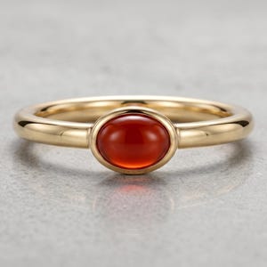 May include: A gold ring featuring a smooth, polished band and a central oval-shaped, deep orange-red cabochon gemstone. The stone is set in a simple gold bezel, creating a clean and uniform appearance for this piece of jewelry.