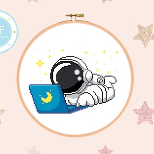 Puede incluir: Patrón de punto de cruz con un astronauta de dibujos animados con un traje espacial blanco, sentado con un portátil, sobre un fondo blanco con estrellas amarillas.