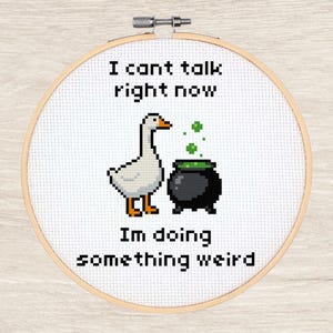 Könnte beinhalten: Kreuzstich-Stickrahmen mit weißem Hintergrund. Es zeigt eine pixelige Gans, einen schwarzen Kessel mit grünen Blasen und den Text "I can't talk right now, I'm doing something weird."