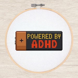 Pode incluir: Bordado em ponto cruz num bastidor de madeira. O desenho apresenta uma bateria pixelizada com o texto "POWERED BY ADHD" em amarelo e vermelho. A bateria é castanha com um sinal de mais.