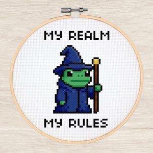 May include: Cross-stitch embroidery in a wooden hoop features a pixelated green frog wizard in a blue robe and hat, holding a staff. The text "MY REALM MY RULES" is stitched above and below the frog.