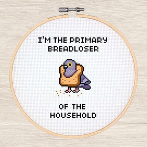 Könnte beinhalten: Ein Kreuzstich-Stickrahmen mit dem Text "I'M THE PRIMARY BREADLOSER OF THE HOUSEHOLD." In der Mitte befindet sich eine pixelige Taube, die ein Brotstück trägt. Die Stickerei befindet sich auf weißem Stoff in einem Holzrahmen.