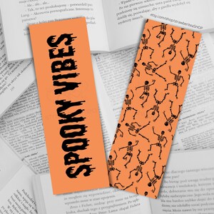 May include: Two Halloween-themed bookmarks. One is orange with the words "SPOOKY VIBES" in black. The other is orange with a pattern of black skeleton figures. Both are rectangular and rest on open books.