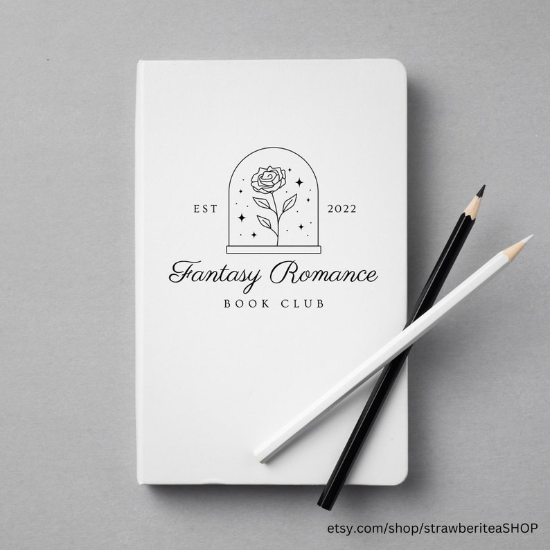 K&ouml;nnte beinhalten: Ein wei&szlig;es Notizbuch mit einem schwarz-wei&szlig;en Logo, auf dem "Fantasy Romance Book Club" steht, sowie dem Text "EST 2022" und einer Strichzeichnung einer Rose in einer Glaskuppel mit Sternen.