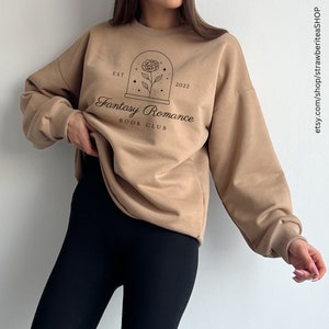 K&ouml;nnte beinhalten: Ein beiger Pullover mit einem schwarzen grafischen Design, auf dem "Fantasy Romance Book Club" mit einer Rose in einer Kuppel und Sternen steht. Der Text "EST 2022" ist ebenfalls im Design enthalten.