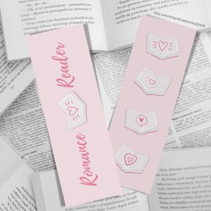 Puede incluir: Marcapáginas rosa y blanco con el texto "Romance Reader" e ilustraciones de libros abiertos con corazones. El marcapáginas está diseñado para los amantes de los libros y presenta un tema romántico.