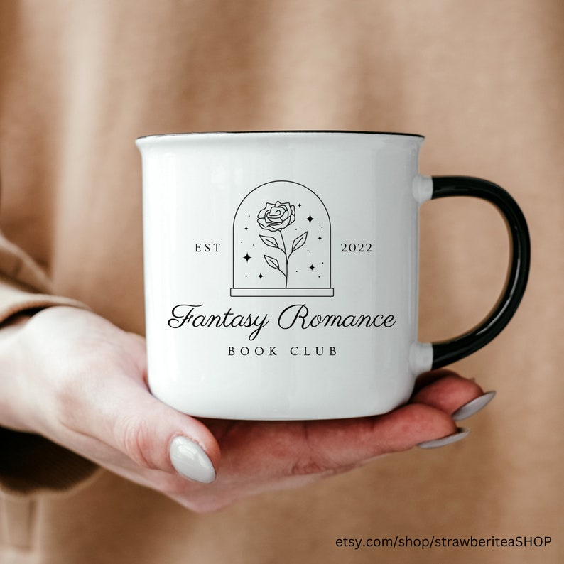 K&ouml;nnte beinhalten: Eine wei&szlig;e Keramiktasse mit schwarzem Henkel. Die Tasse hat einen schwarzen Rand und ein Design einer Rose in einer Kuppel mit Sternen. Der Text "EST 2022 Fantasy Romance Book Club" ist auf die Tasse gedruckt.