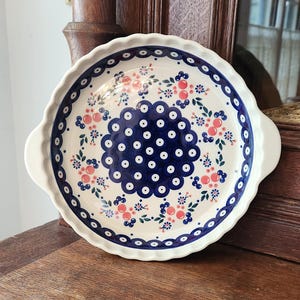 May include: Round ceramic serving dish with a scalloped edge. The dish features a white base with a blue and pink floral pattern. It has two small handles and is displayed on a wooden surface.