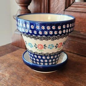 Op de afbeelding: Een keramische bloempot met bijpassende schotel. De pot is wit met een blauwe rand en is versierd met blauwe, rode en witte bloempatronen. De pot staat op een houten oppervlak.