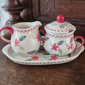 Könnte beinhalten: Ein Milchkännchen und eine Zuckerdose mit Deckel auf einem ovalen Tablett. Die Keramik ist weiß mit einem Muster aus rosa Blumen sowie blauen und grünen Blättern. Jedes Teil hat einen roten Henkel, einen roten Deckelknauf und rote Punkte am Rand.