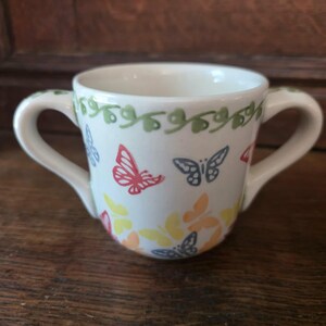 May include: A white ceramic cup with two handles, adorned with colourful butterflies and green vine details. The butterflies are in red, blue, yellow, and orange. The cup is placed on a wooden surface.