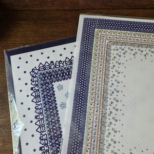 May include: Two rectangular paper sheets in clear plastic sleeves. The left sheet features a dark blue border with stars, snowmen, and trees. The right sheet displays a multi-layered border of small blue flowers, dots, and geometric patterns.