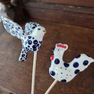 May include: Two ceramic garden stakes featuring a white bird with blue polka dots and a white rooster with blue polka dots, a red comb, and a red wattle. Each figure is mounted on a thin, light-colored wooden stick for placement in soil.