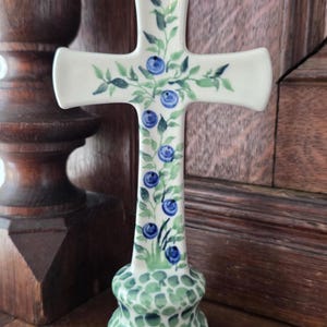 May include: A ceramic cross standing on a rounded base, featuring a hand-painted pattern of green leaves and dark blue berries along the vertical stem and horizontal arms, set against a smooth, off-white glazed surface with a green dotted base.