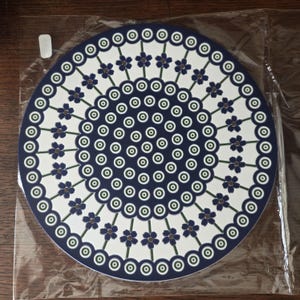 May include: A circular plate featuring a dark blue center filled with concentric rings of small white and green circles. The outer white border displays a repeating pattern of dark blue five-petaled flowers on green stems, framed by a dark blue edge.