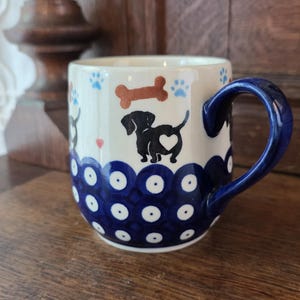 May include: A ceramic mug featuring a cream-colored background with a pattern of black dachshund silhouettes, each with a white heart on its side. The design includes brown dog bones, small red hearts, and blue paw prints, with a blue polka dot base.
