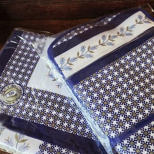 Puede incluir: Un conjunto de dos artículos textiles plegados en un embalaje de plástico transparente. La tela presenta un patrón geométrico azul oscuro y blanco, un borde blanco con un estampado de enredadera floral azul y un logo circular con una B.