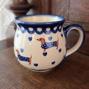 Puede incluir: Una taza de cerámica de color crema con un patrón pintado a mano de perros salchicha marrones con suéteres azules de lunares, intercalados con pequeños corazones azules. El borde tiene un diseño festoneado azul y un asa curva.