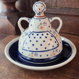 Könnte beinhalten: Eine Keramik-Butterdose mit einem cremefarbenen, gewölbten Deckel mit zwei Griffen, verziert mit blauen Punkten, einem zentralen Schürzenmotiv sowie kleinen orangefarbenen und blauen Blumen. Der Deckel hat einen runden Knauf und steht auf einem dunkelblauen Teller.