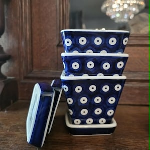 May include: A stack of four blue and white ceramic square dishes with a matching lid. The dishes have a dark blue background with white dot patterns. The lid is propped up next to the stack. The dishes are on a wooden surface.
