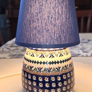 May include: A table lamp with a dark blue fabric drum shade and a ceramic base. The base features a rounded shape with a white background and a repeating pattern of blue, green, and orange floral and geometric motifs across its surface.